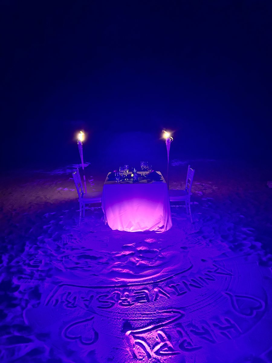 A private candlelit dinner for two set up on the sand at night, with HAPPY ANNIVERSARY written in the sand around the table