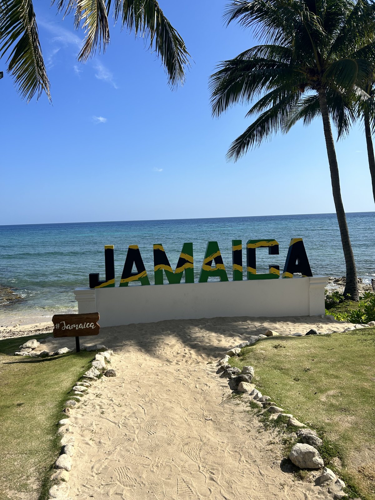 The JAMAICA beach sign at Hyatt Zilara Rose Hall in Montego Bay, palm trees and turquoise Caribbean water behind it